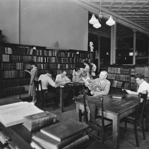 black and white image of patrons gather in the library reading at tables.