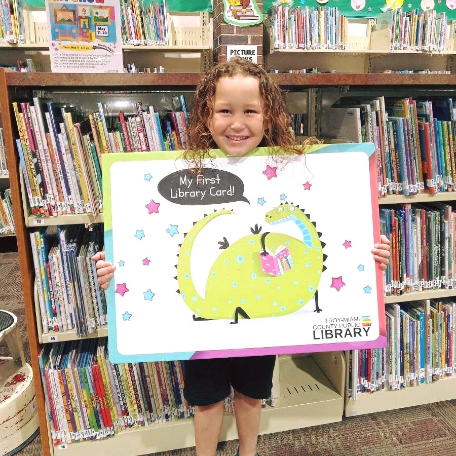 "a little boy stands in front of a book shelf holding a sign that says my first library card"